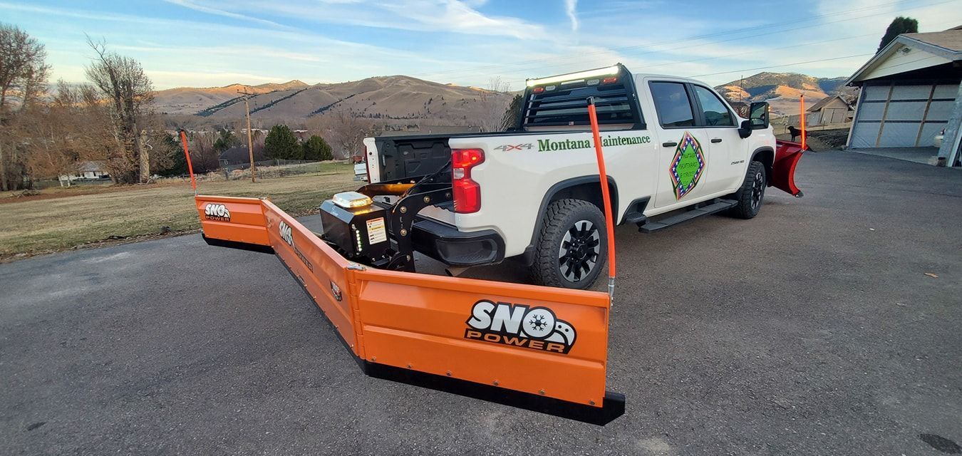 White pickup truck with snowplow parked on asphalt. Mountains and house in the background.