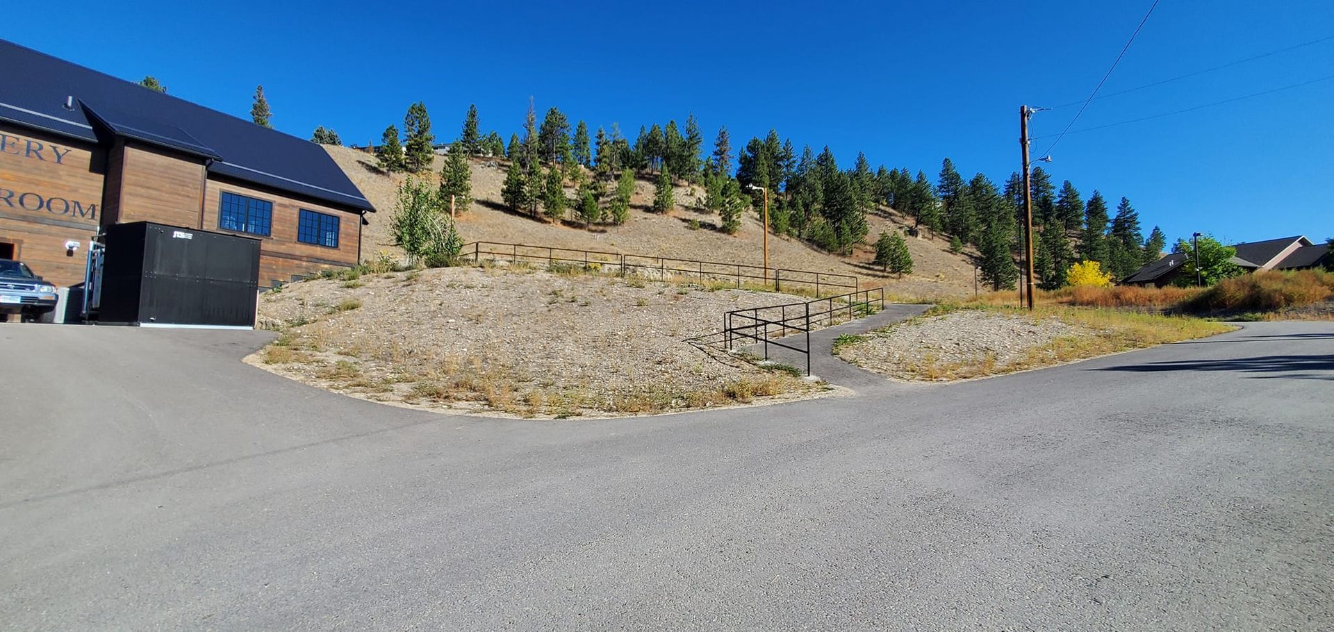 Road leads to a building with solar panels. A dry hillside with trees is behind it. Bright blue sky.