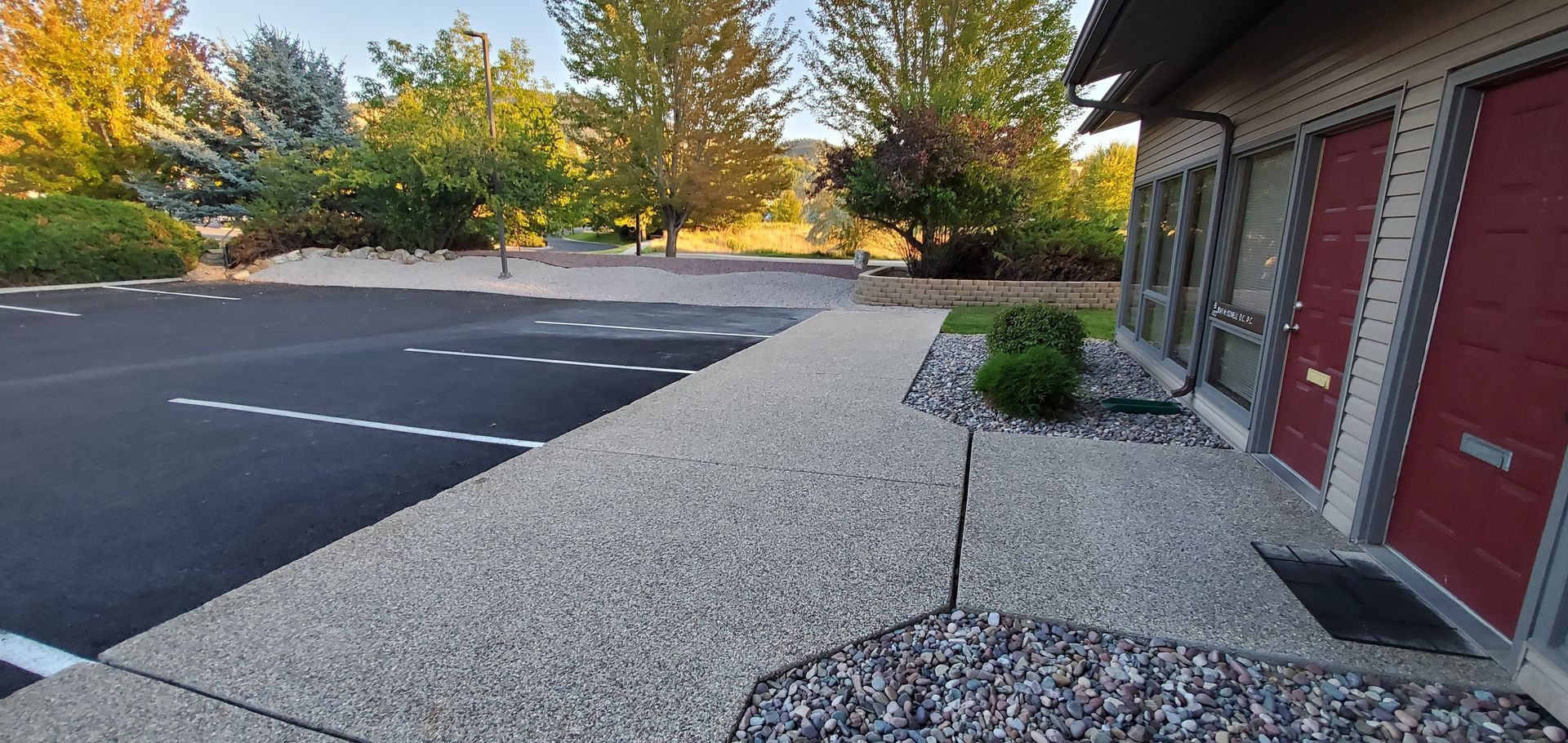 A paved walkway with a cracked surface, leading to a building with red doors. A parking lot is to the left.