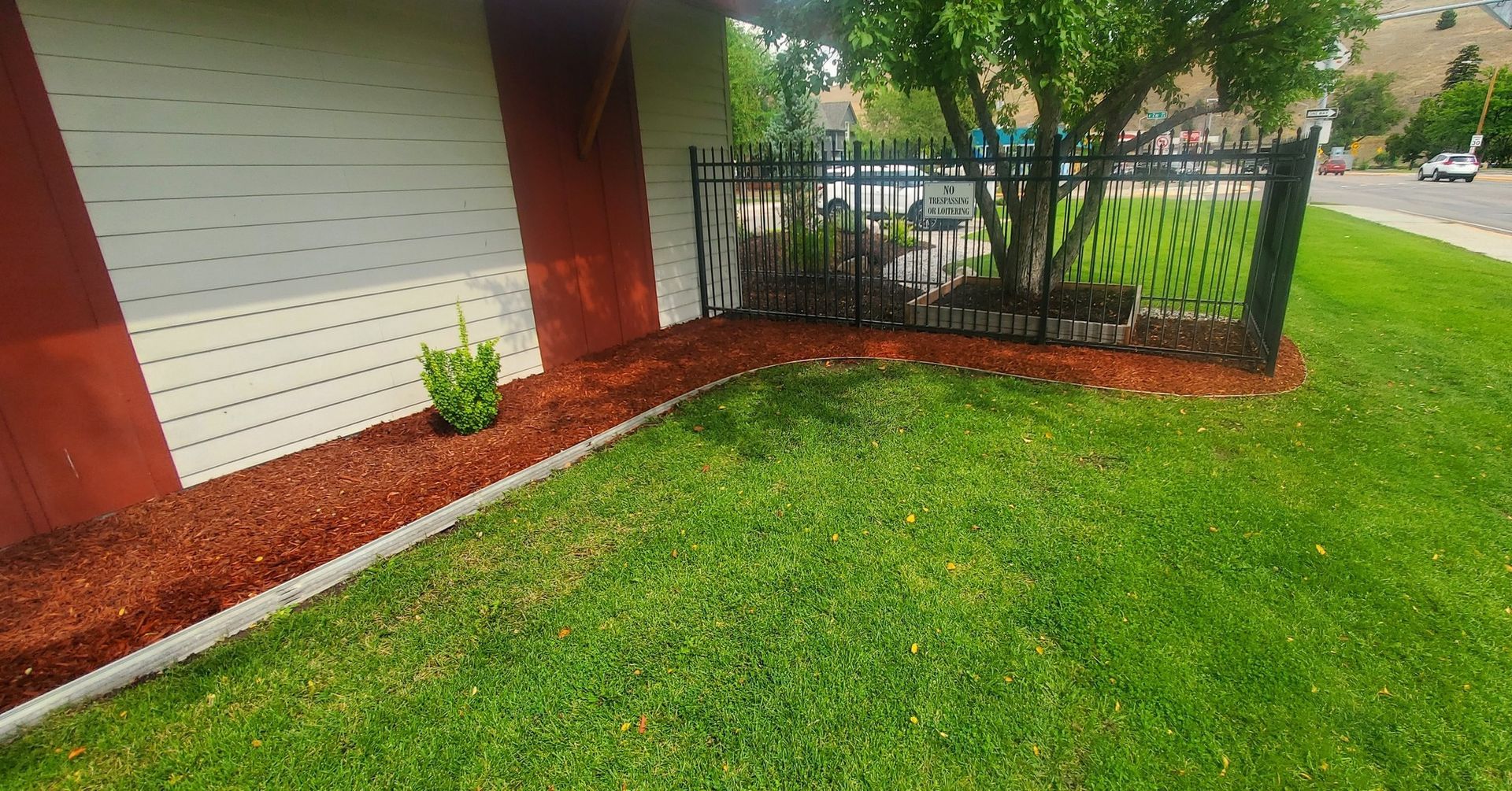Red-painted building with green lawn, mulch bed, black fence, and a tree. Sunny outdoor setting.