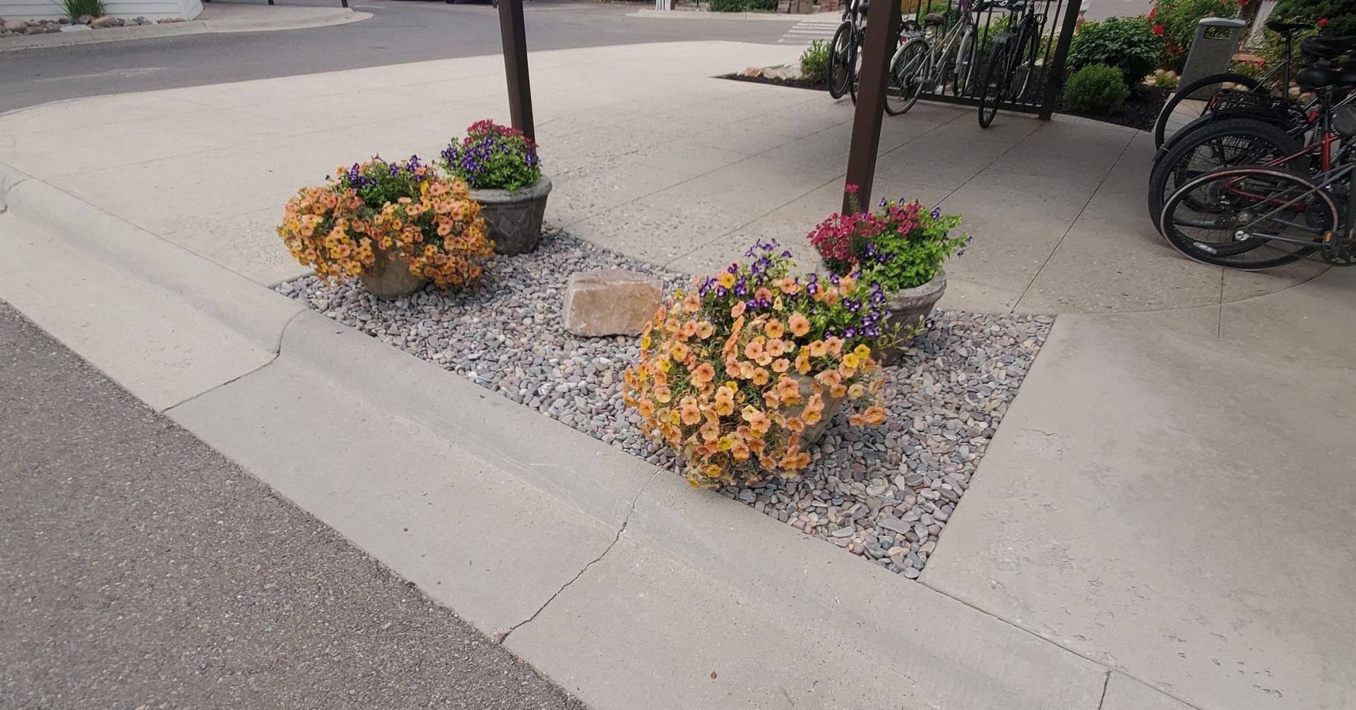 Flowers in pots and gravel landscaping bordering a sidewalk, next to a street with parked bicycles.
