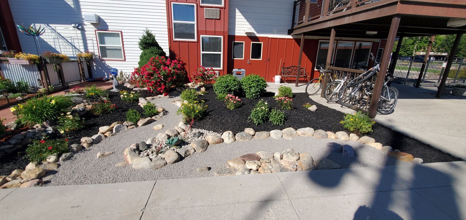 A colorful garden bed with flowers, mulch, and rocks in front of a red and white building.