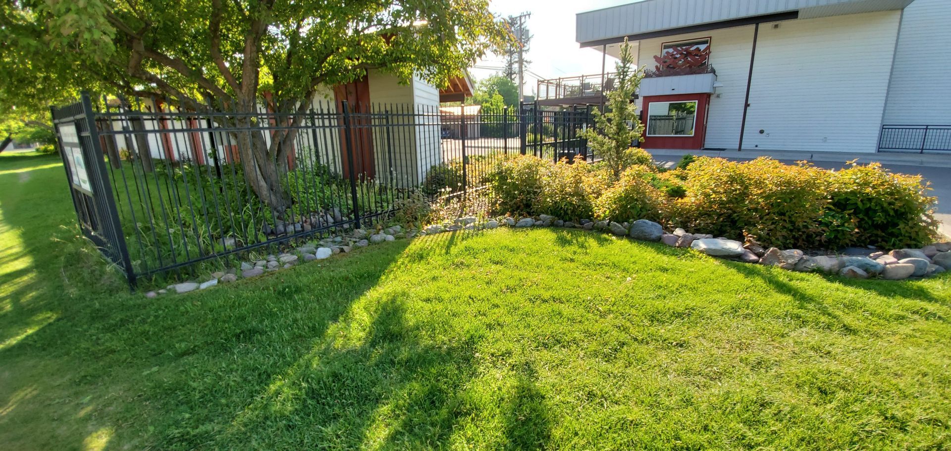 Green lawn, flowerbeds with rocks, black fence, and a white building with a dark roof.