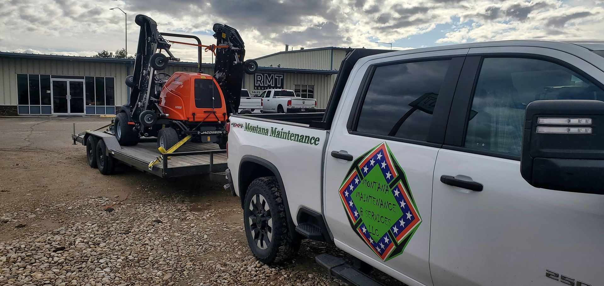 A white truck with a trailer carrying an orange construction machine in a gravel parking lot.