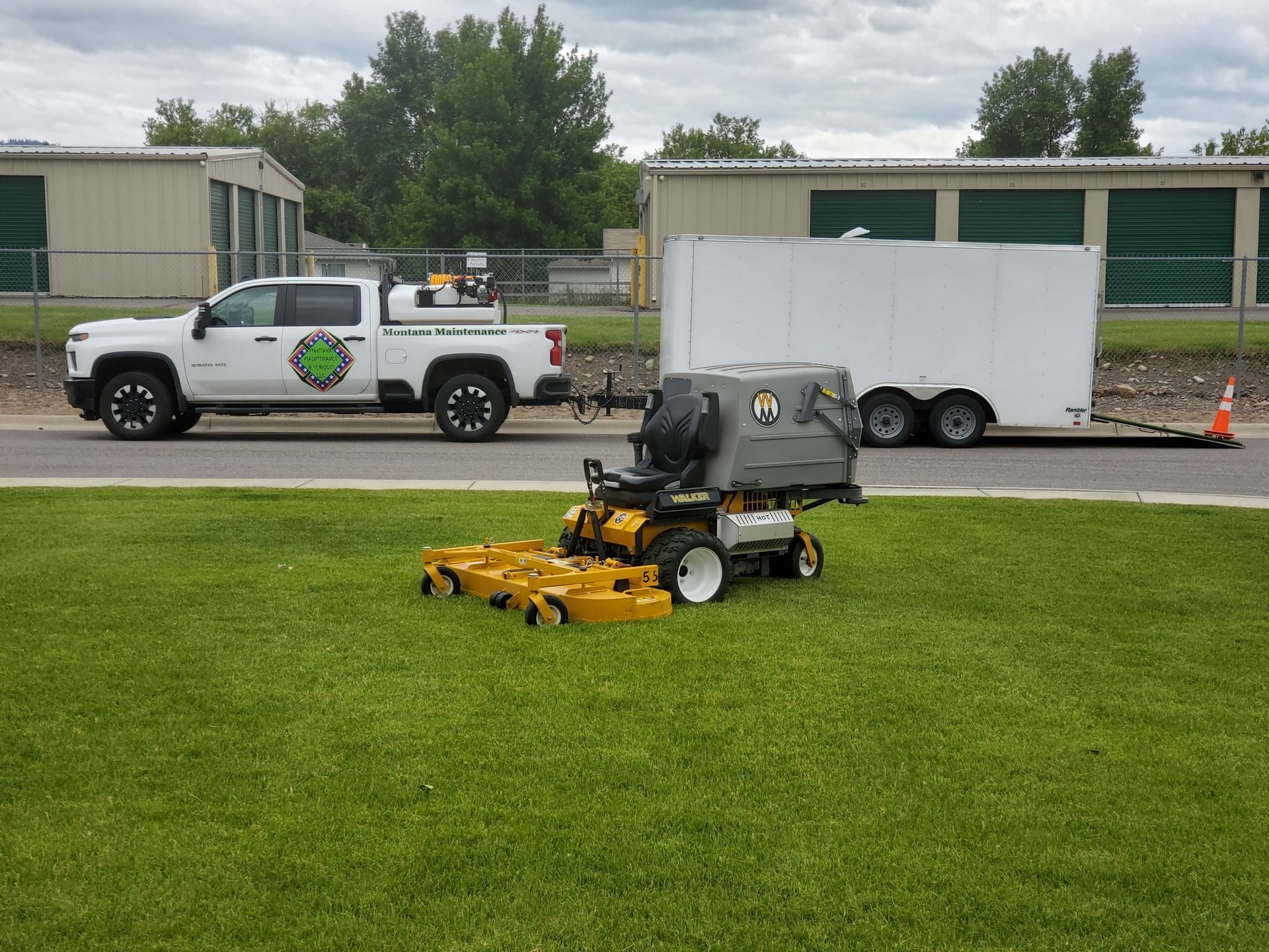 A lawn care truck towing a trailer, with a riding mower on grass, parked near storage units.
