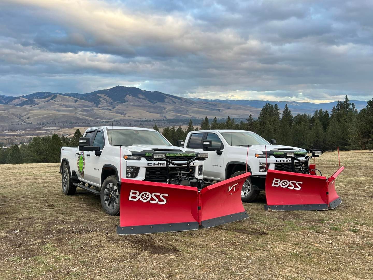 Two white pickup trucks with red snow plows parked on a hill with mountains in the background.