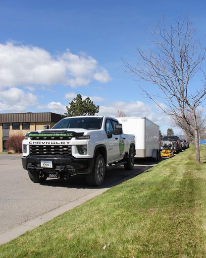 White Chevy truck pulling a trailer, parked on a road next to a grassy area with a building in the background.