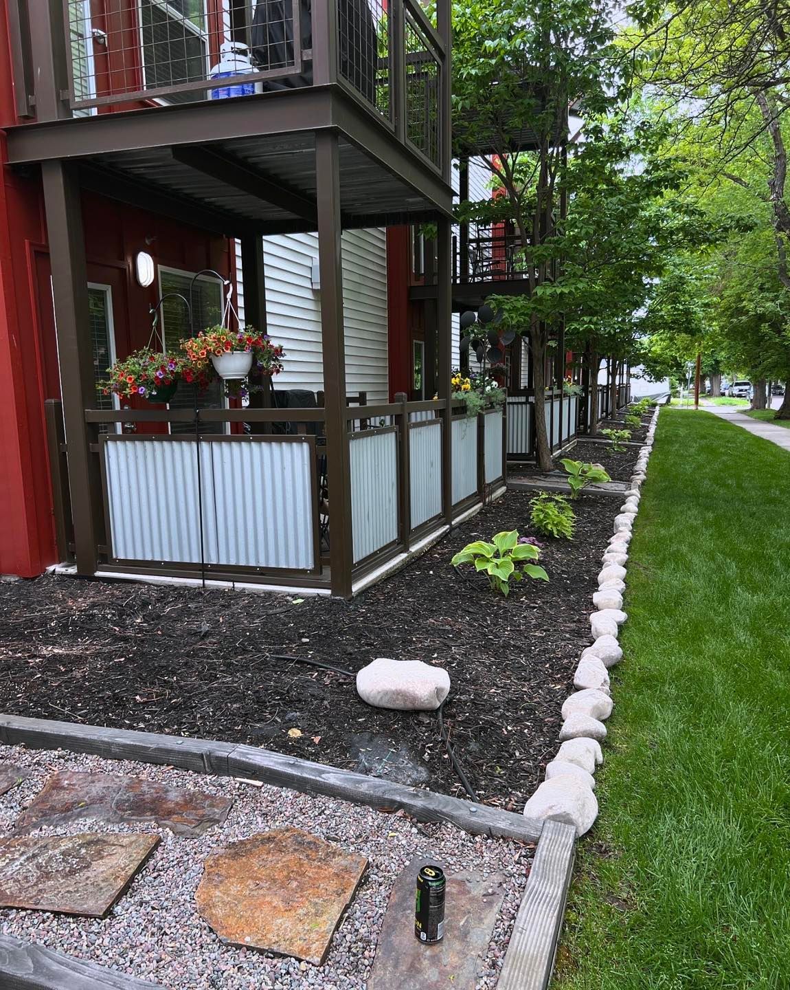 Red building with metal balconies, planter boxes, and corrugated metal fence, lined with plants and rocks.