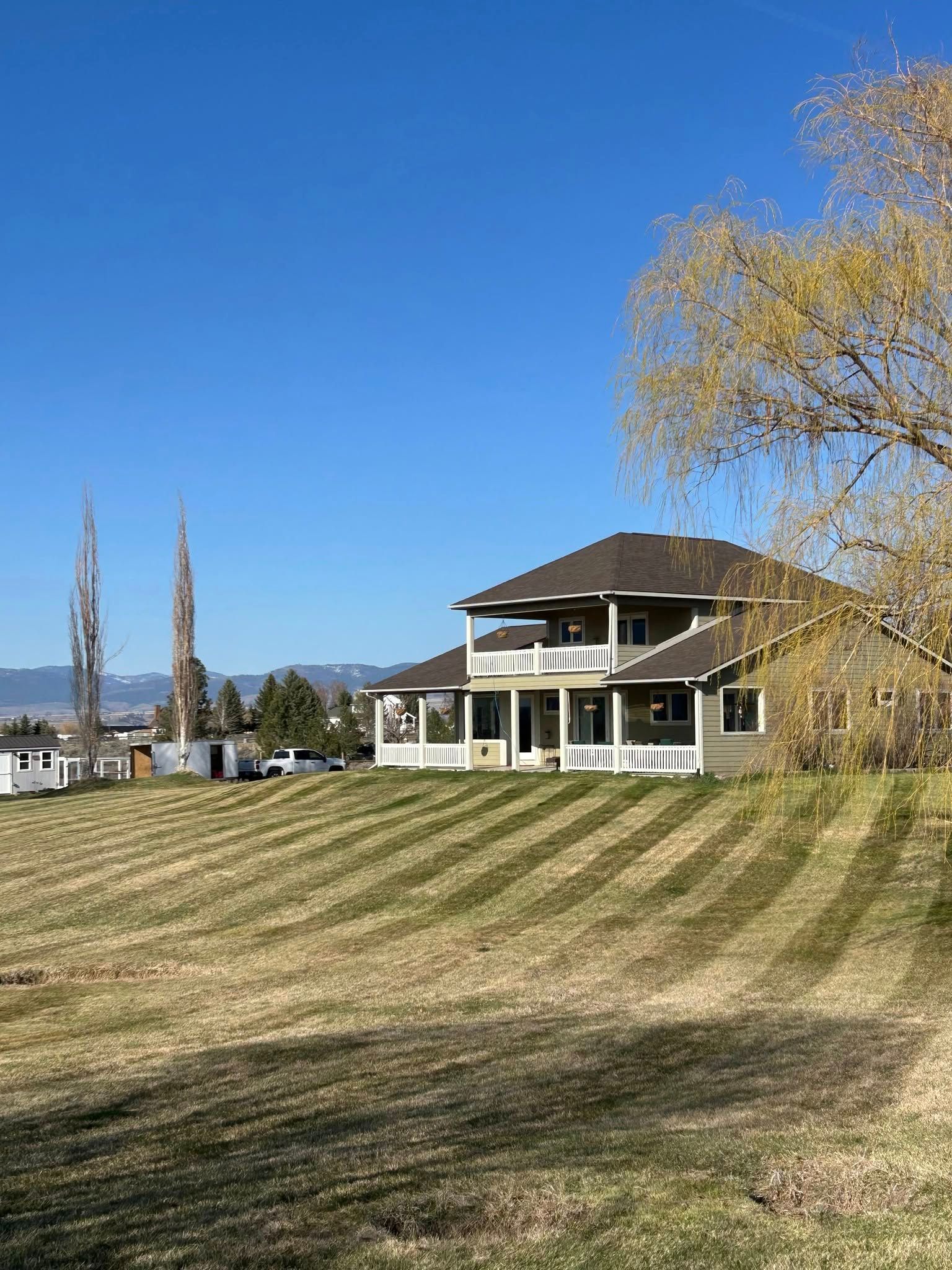Two-story house with a wraparound porch, behind a mowed lawn and tall bare trees, against a bright blue sky.