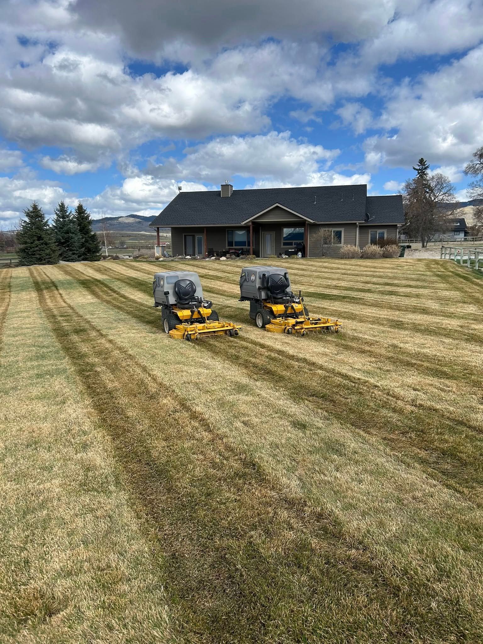 Two riding lawn mowers cutting stripes in a grassy lawn in front of a house on a sunny day.