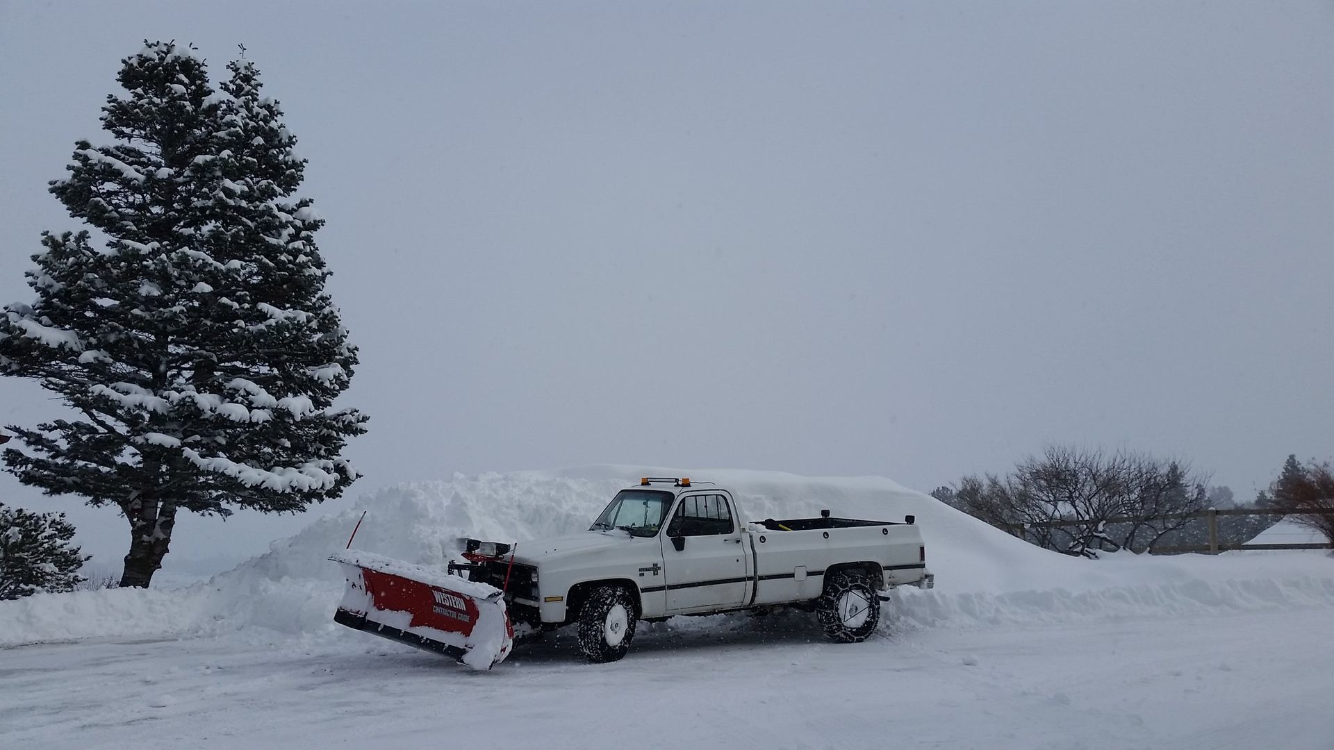 White pickup truck plowing snow on a snow-covered road. A large snow pile and tree are in the background.