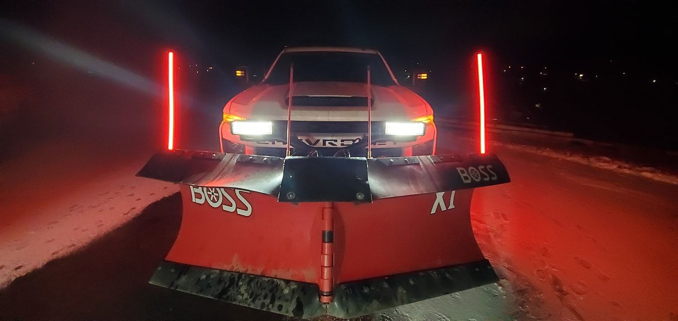 A snow plow on a truck at night, red lights glowing, clearing a snow-covered road.