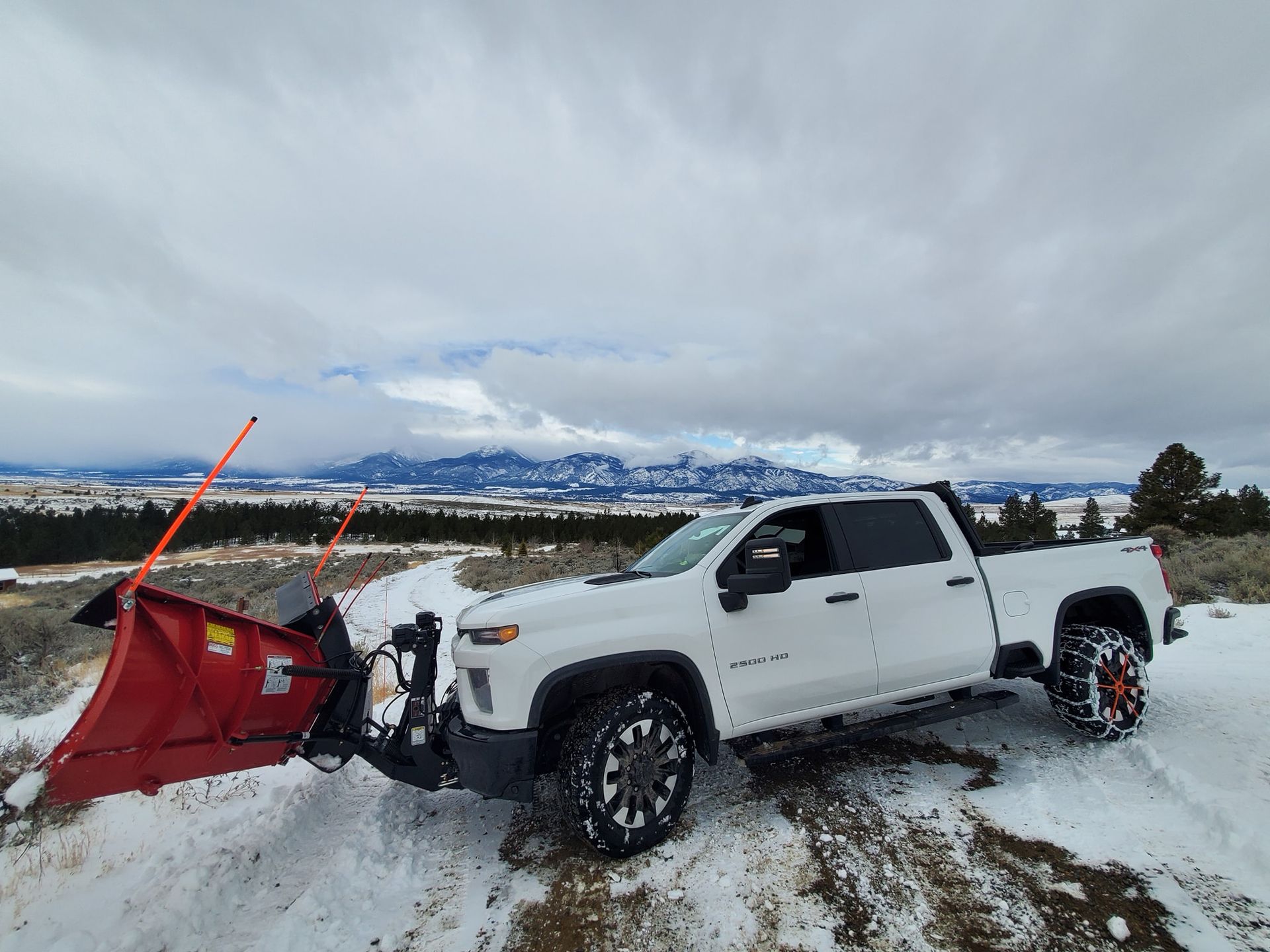 White truck with snow plow on a snowy road with mountains in the background. Overcast sky.