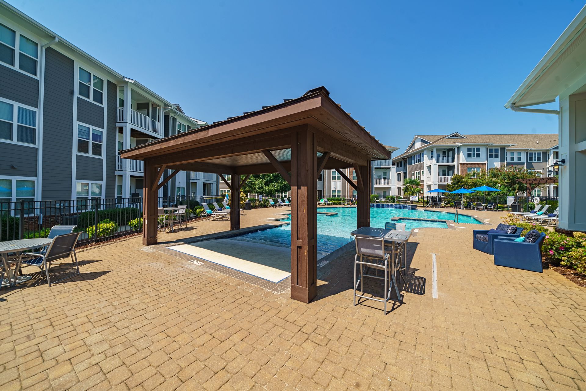 Poolside patio with a wooden gazebo, tables, and chairs; apartment buildings in the background under a blue sky at Willows at Fort Mill, offers apartments for rent near Downtown Fort Mill, SC.