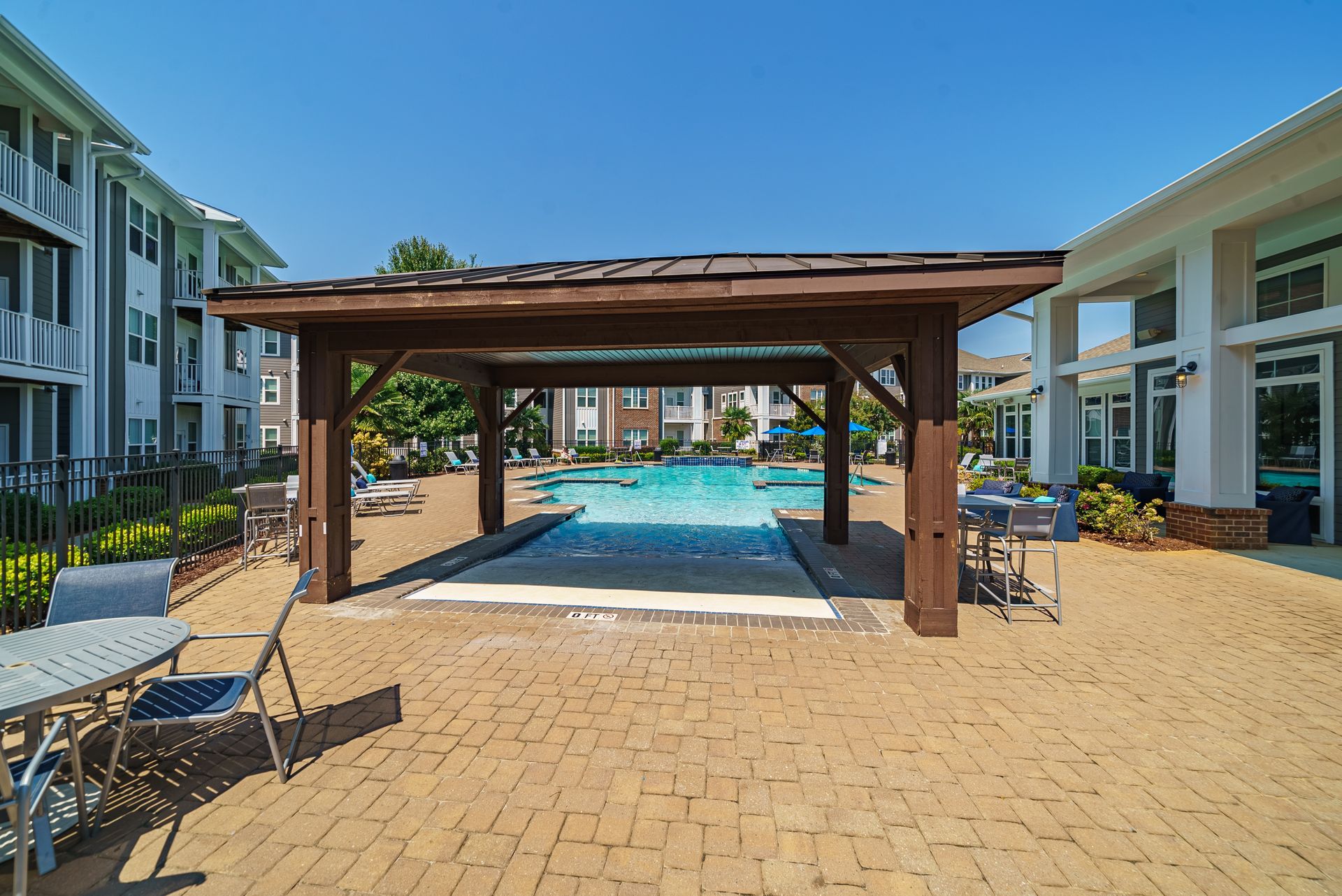 Poolside with pergola, tables, chairs, and buildings at Willows at Fort Mill, offers apartments for rent near Downtown Fort Mill, SC. Blue sky, water, and brick ground.
