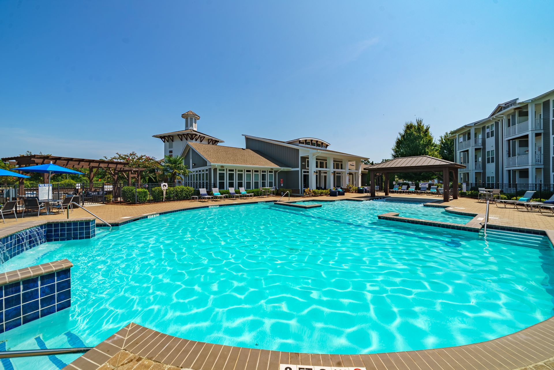 Swimming pool and clubhouse on a sunny day at Willows at Fort Mill, offers 1, 2, and 3 bedroom apartments for rent in Fort Mill, SC. Blue water, clear sky, buildings in the background.