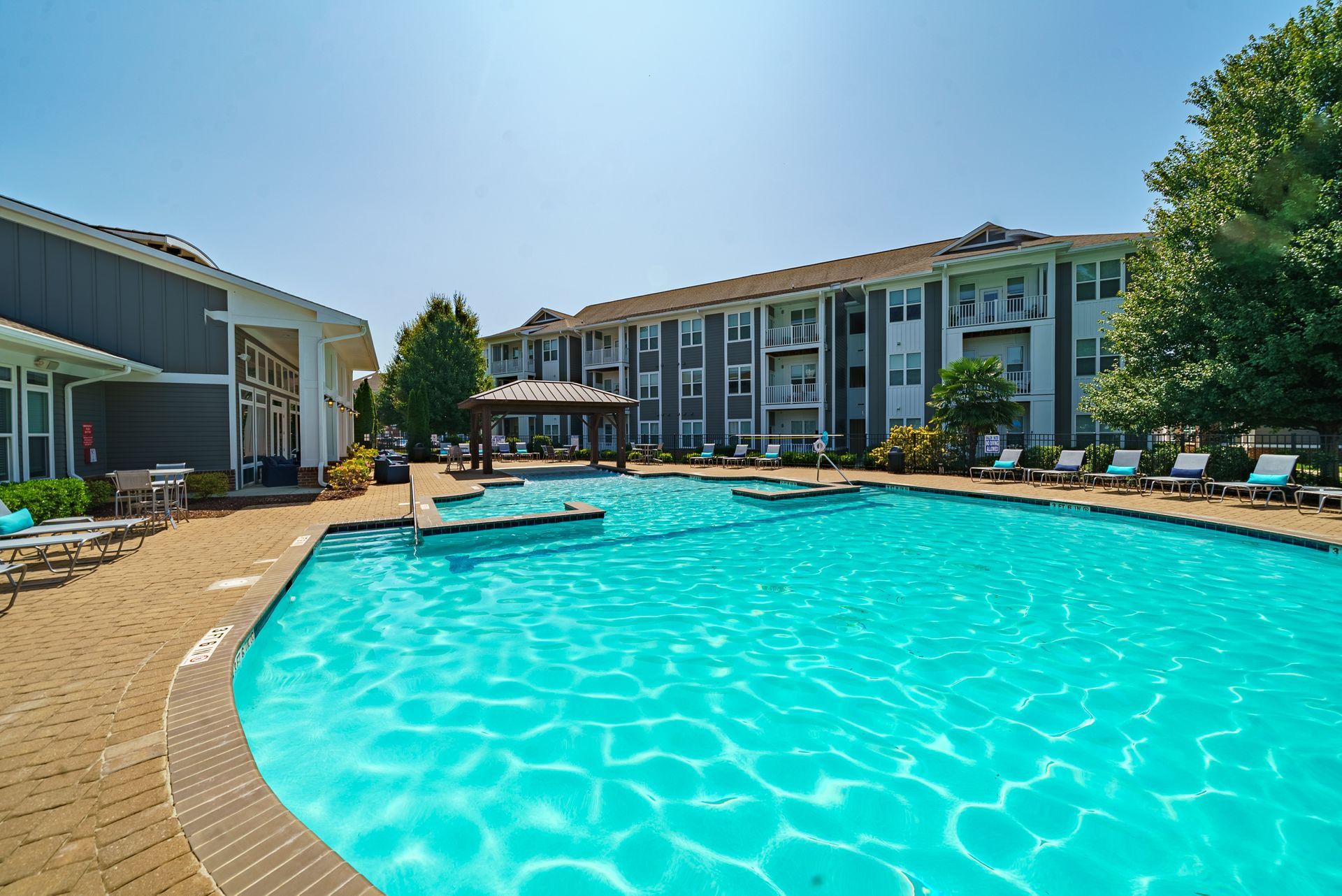 Pool area with turquoise water, lounge chairs, and a gazebo, next to apartment buildings at Willows at Fort Mill, offers 1, 2, and 3 bedroom apartments for rent in Fort Mill, SC.