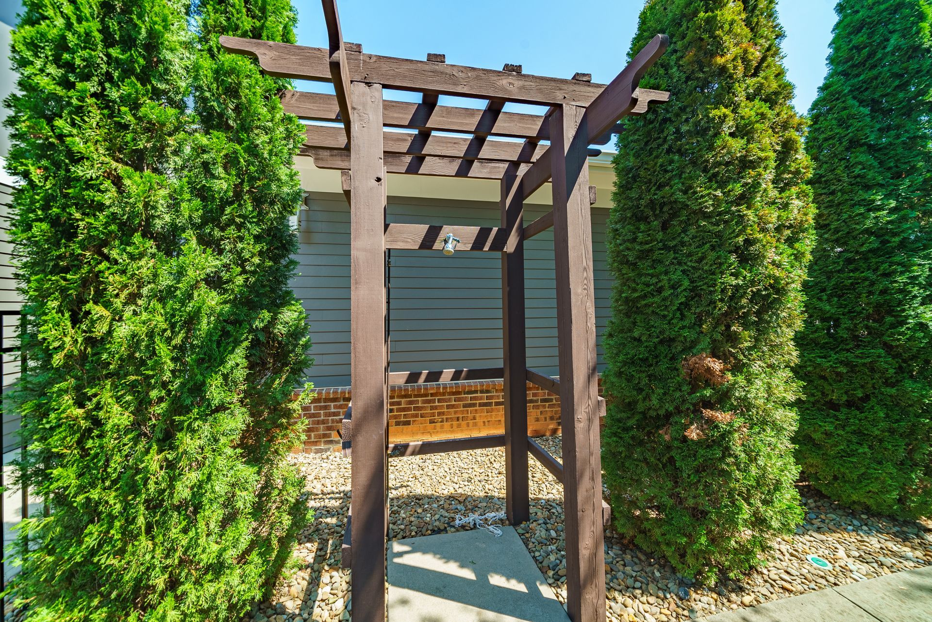 Wooden arbor entrance with tall green trees, gravel ground, and light blue building background at Willows at Fort Mill, offers 1,2, and 3 bedroom apartments near Kingsley Town Center in Fort Mill, SC.