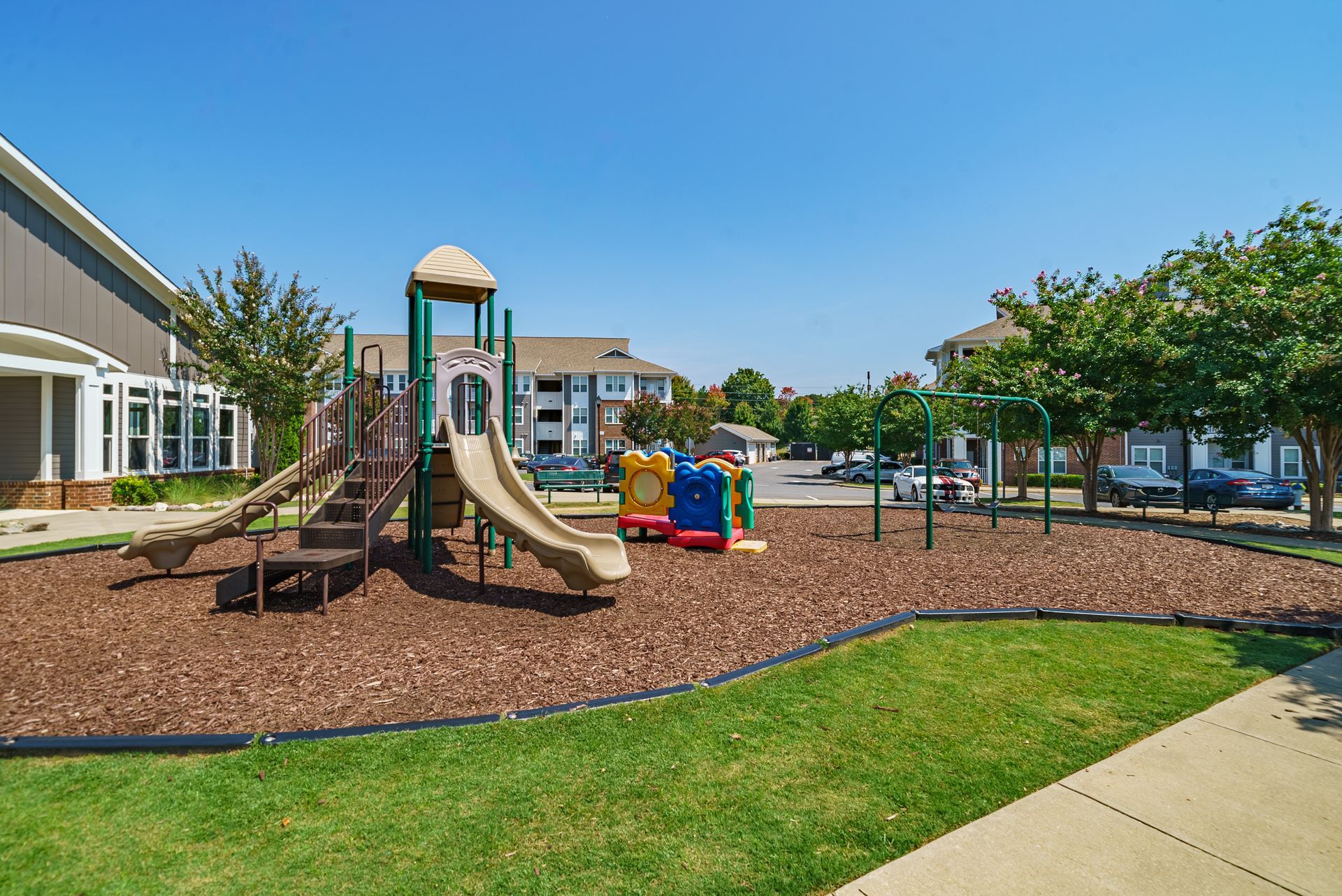 Playground with slide, swings, and mulch on green lawn, with apartment buildings in background at Willows at Fort Mill, offers apartments for rent near Kingsley Town Center in Fort Mill, SC.