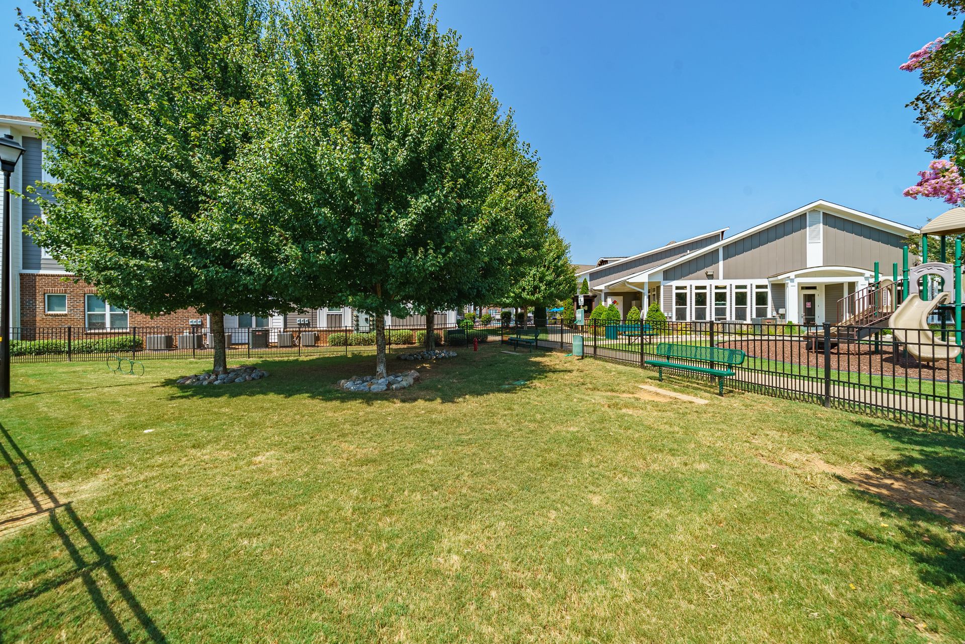 Grassy area with trees, a fence, and two-story buildings under a clear blue sky at Willows at Fort Mill, offers 1,2, and 3 bedroom apartments near Uptown Charlotte in Fort Mill, SC.