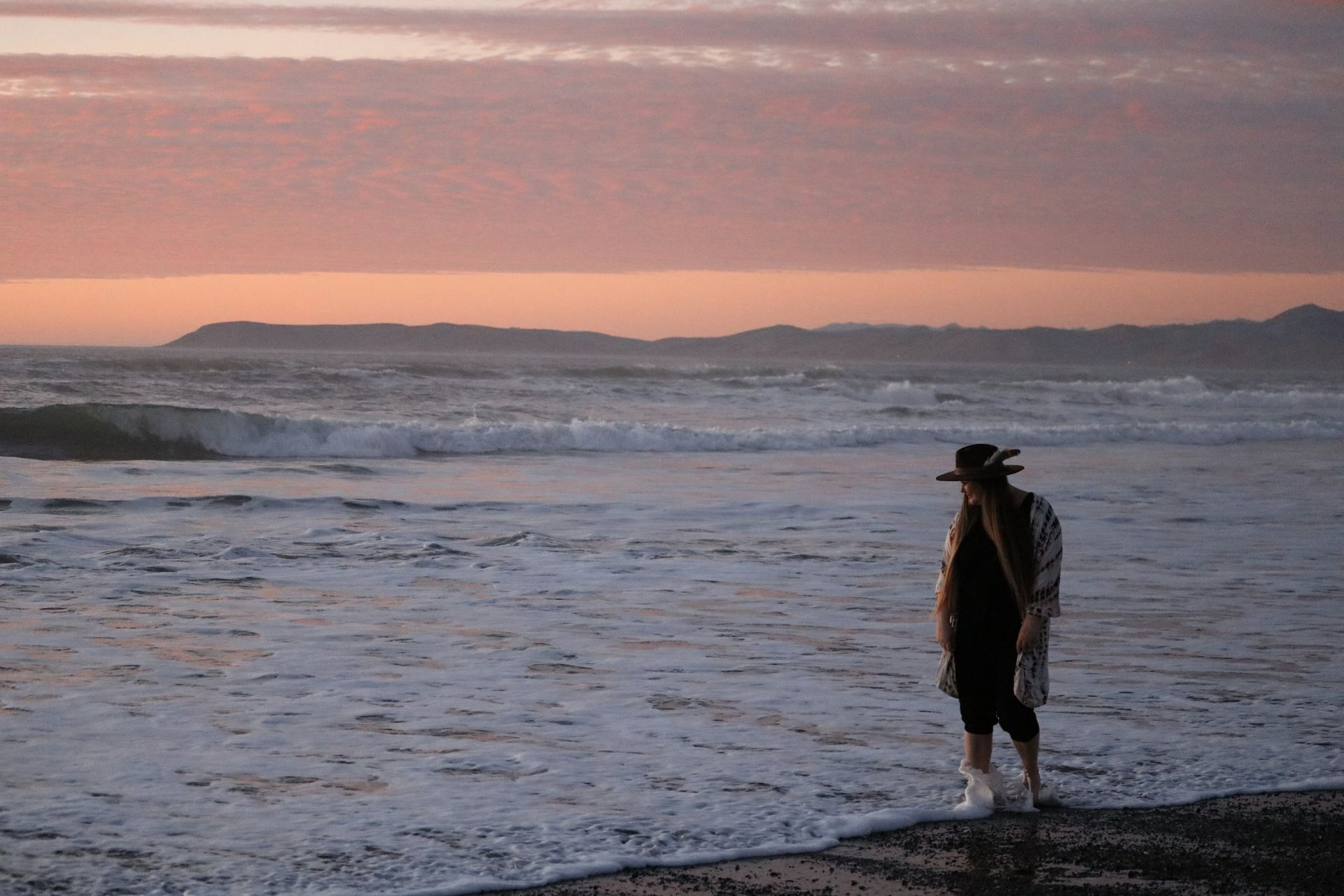 A woman is standing on a beach near the ocean at sunset.