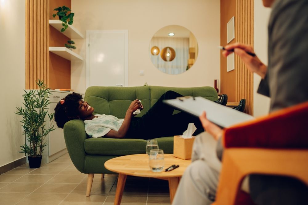 A woman is laying on a green couch while a man holds a clipboard.