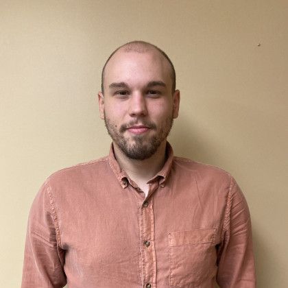 A bald man with a beard wearing a pink shirt is standing in front of a yellow wall.
