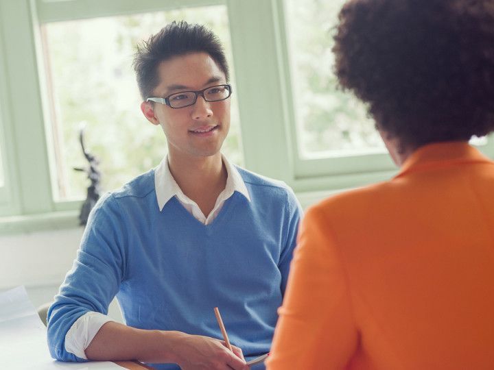 A man in a blue sweater is sitting at a table talking to a woman in an orange jacket.