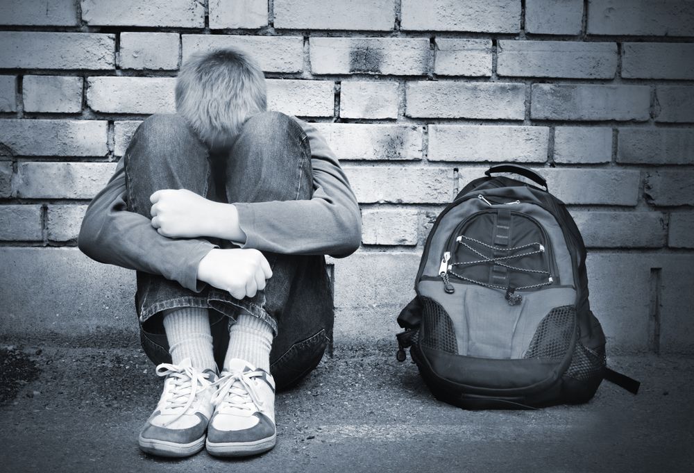 A boy is sitting on the ground with his head down next to a backpack.