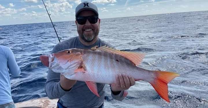 A smiling person wearing a hat and sunglasses holds a large hogfish on a boat on the ocean.