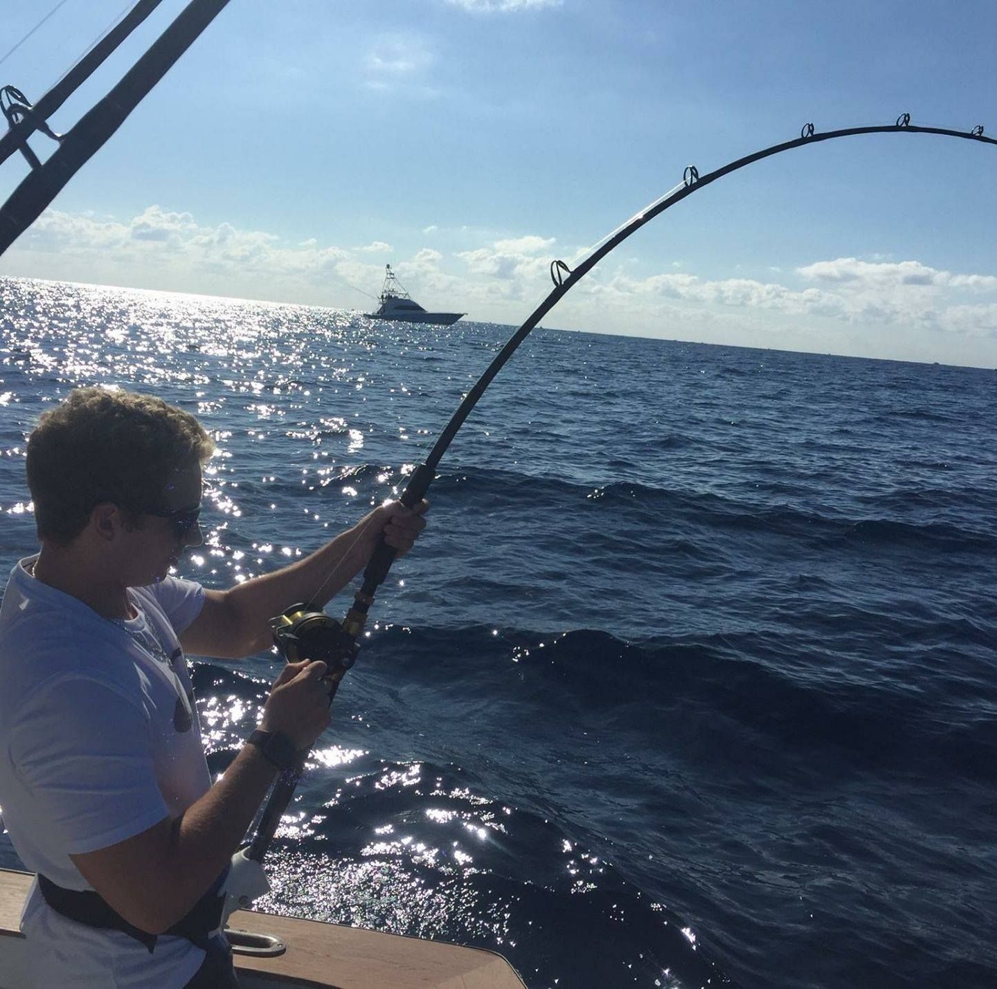 A person fishing on a boat on the ocean with a bent fishing rod, another boat visible in the distance.