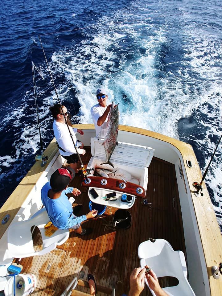 Three people on a boat deck, two standing near a cooler with a large fish while one sits nearby on the wooden floor.