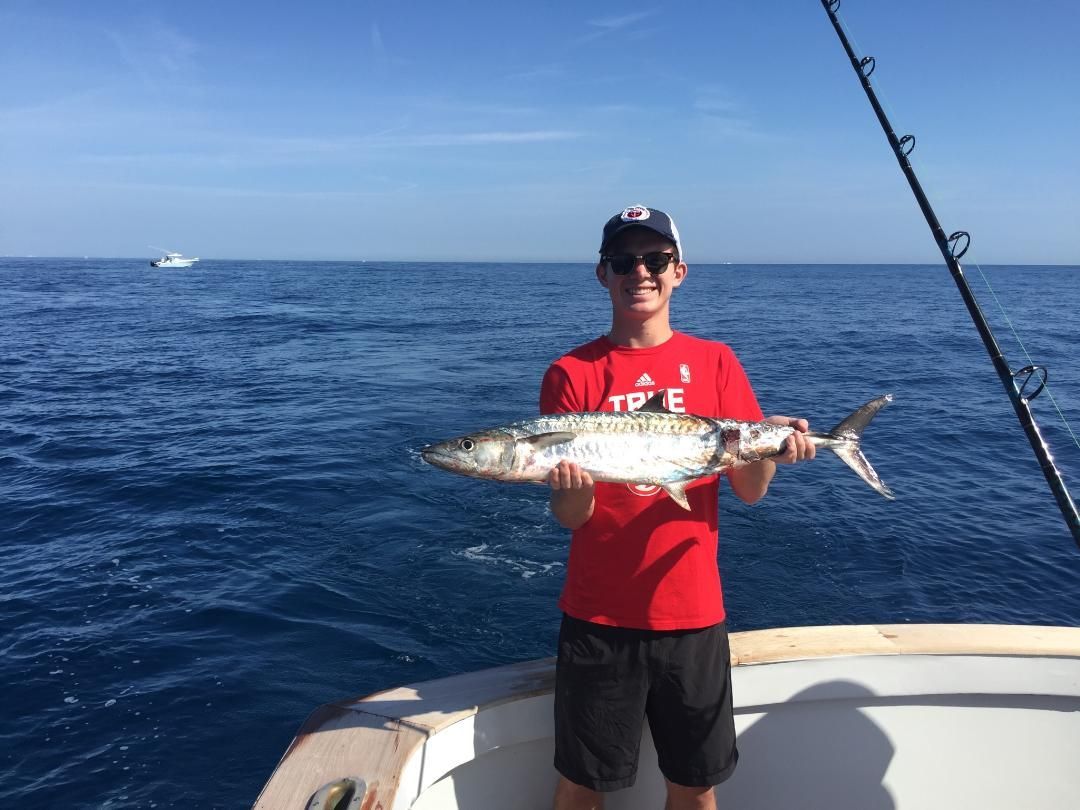 A person in a red shirt and cap holds a fish while standing on a boat in the open ocean under a clear blue sky.