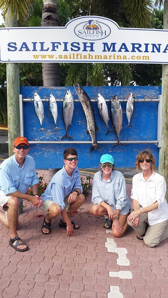 Four people kneel before a rack of caught fish at Sailfish Marina, with a large barracuda centered among tuna.