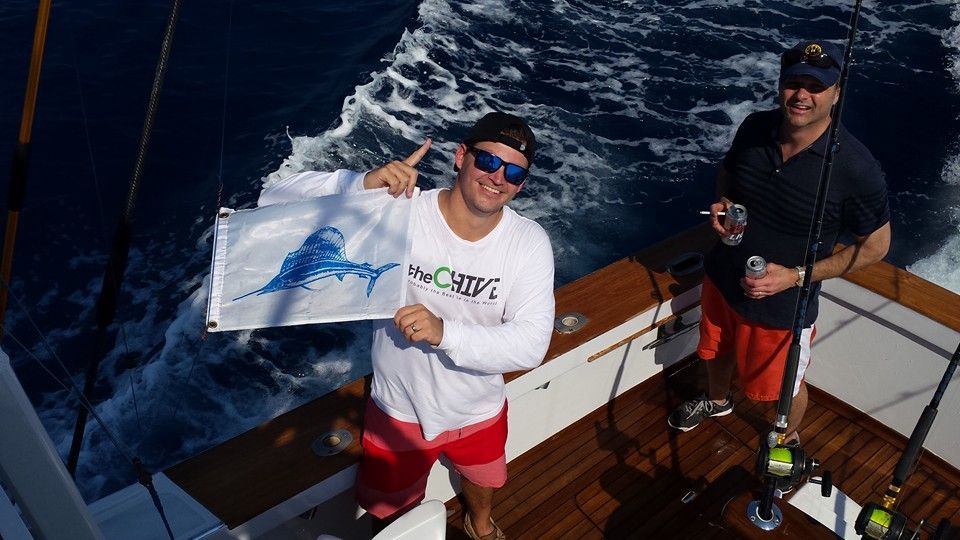Two people on a boat deck, one holding a white flag with a blue marlin graphic while the other stands nearby.