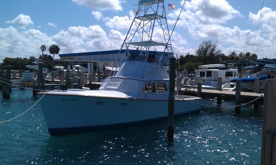 A white fishing boat with a tall tuna tower docked at a marina under a bright, partly cloudy blue sky.