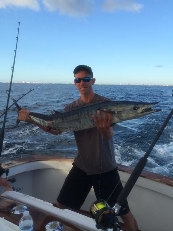 A person holds a large, striped wahoo fish while standing on the deck of a boat on the ocean under a clear blue sky.