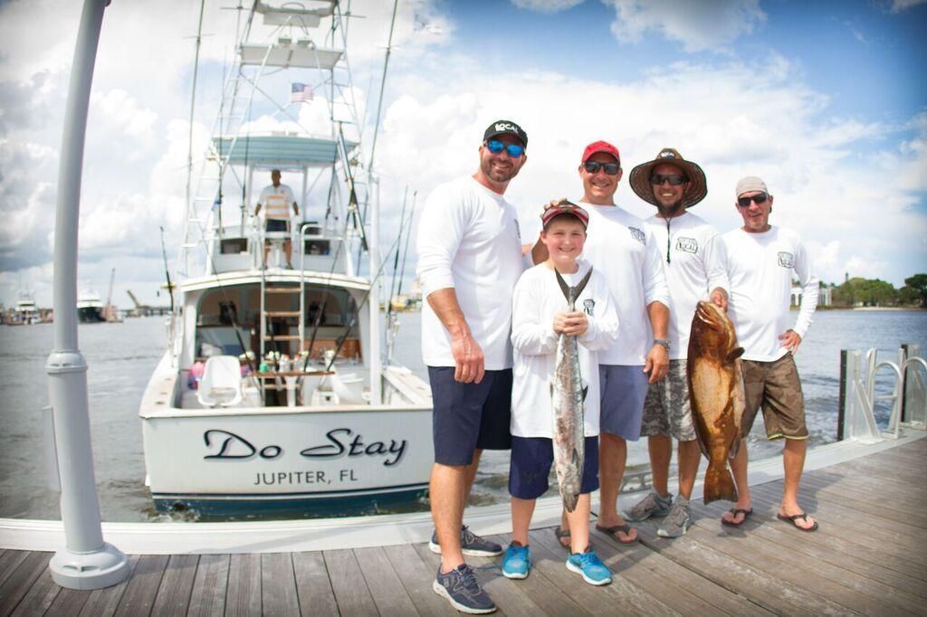 A group of five people stand on a dock holding fish in front of a fishing boat named Do Stay.