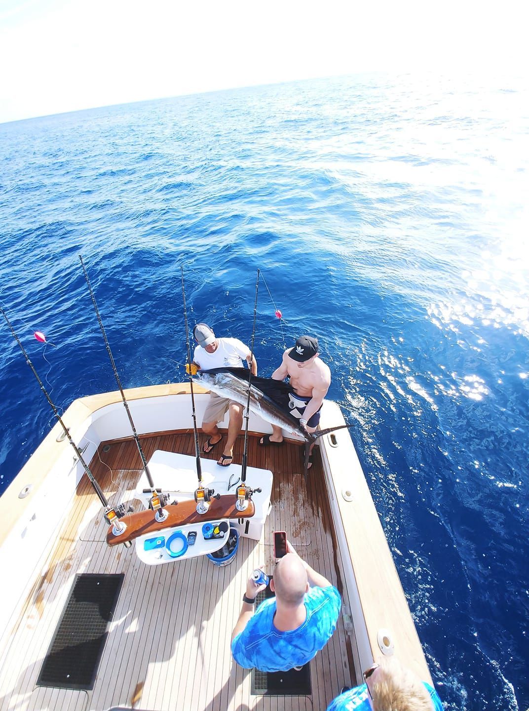 Three people on a boat deck, two standing at the stern holding a large, caught fish while one photographs them.