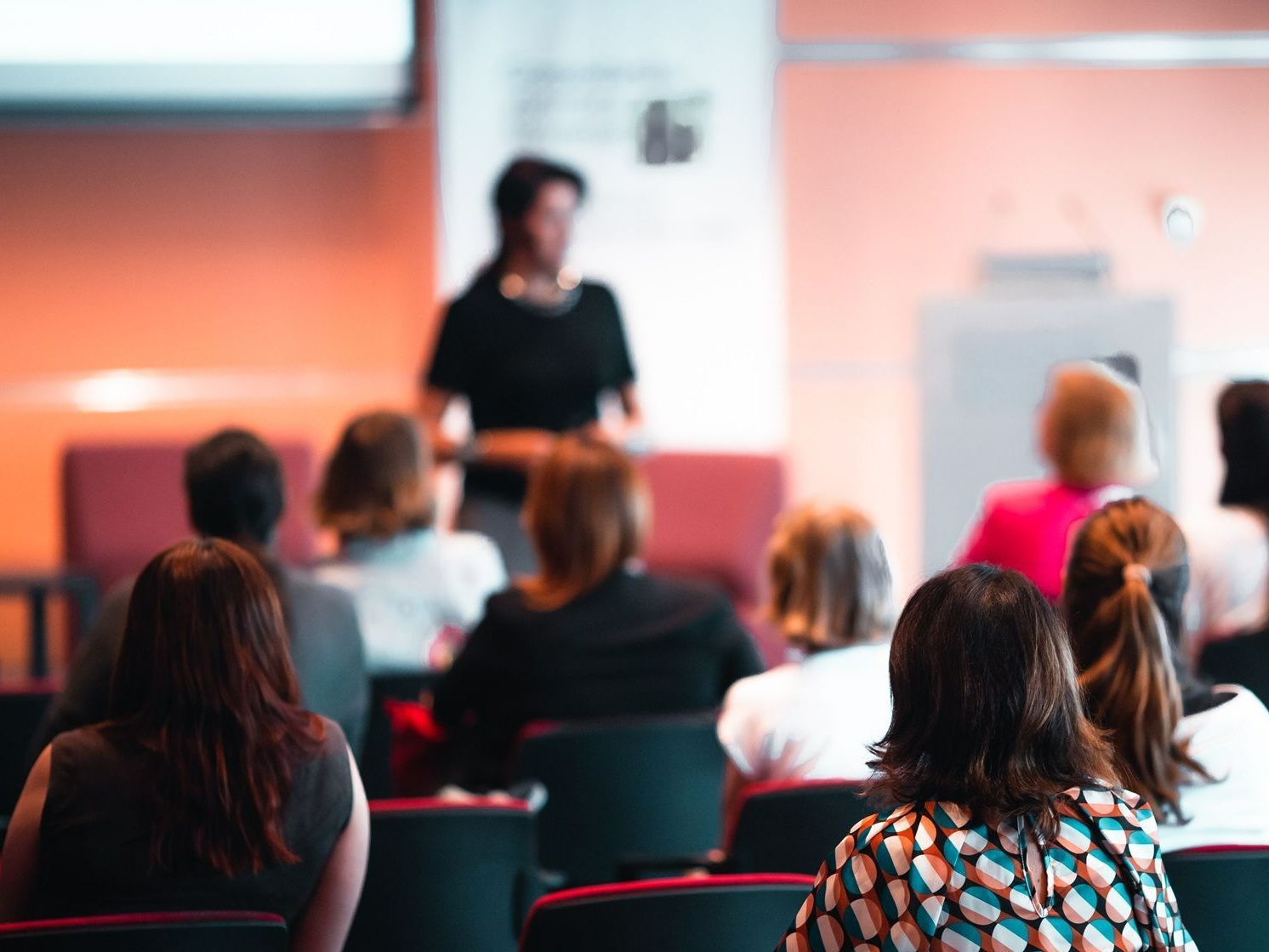 A group of people are sitting in a lecture hall watching a woman give a presentation.