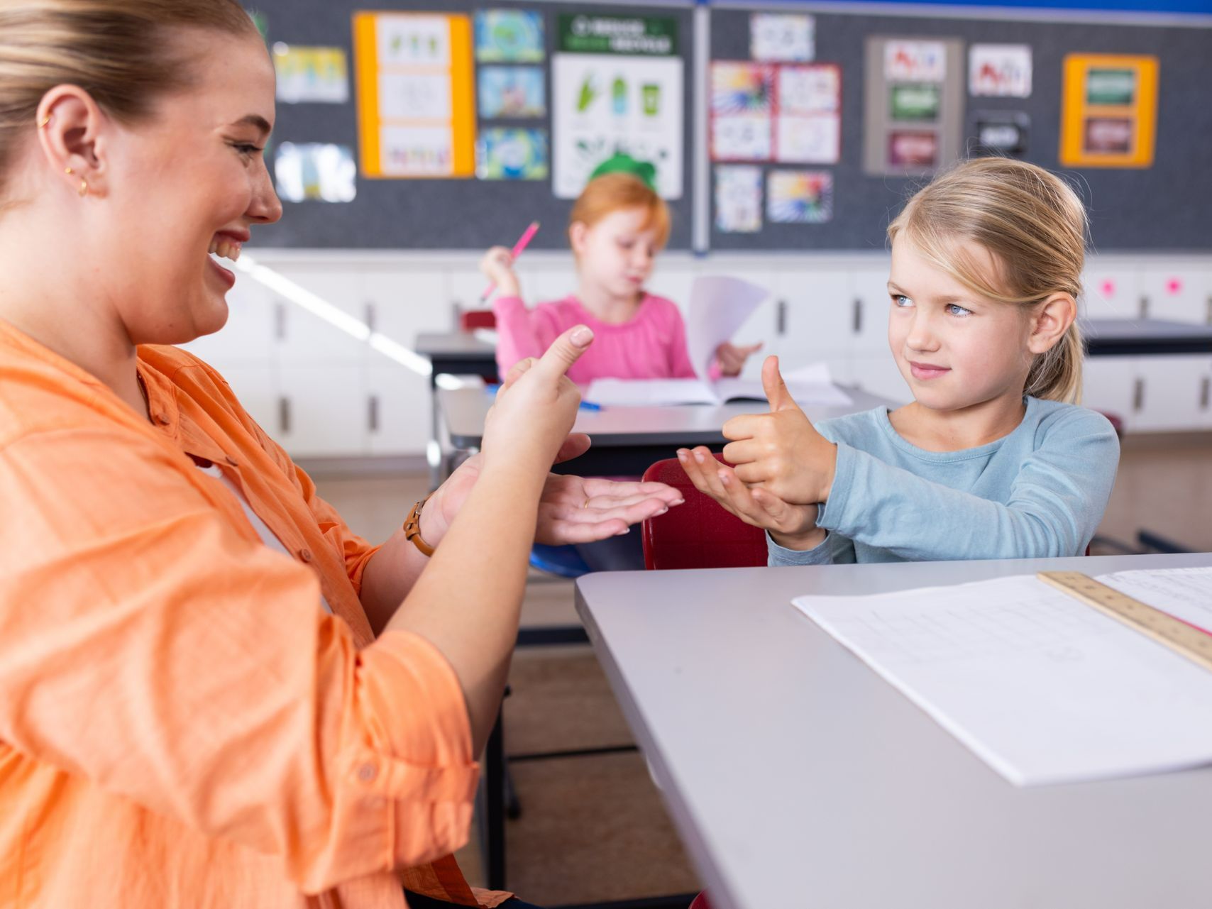 A teacher is giving a thumbs up to a young girl in a classroom.