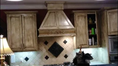 Kitchen with distressed cream-colored cabinets and range hood, dark backsplash, and overhead lighting.