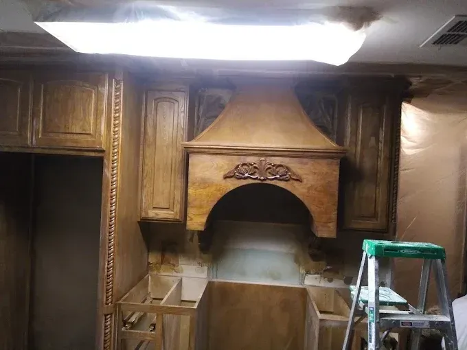 Kitchen cabinets and range hood, stained wood with decorative carvings, under construction with ladder.