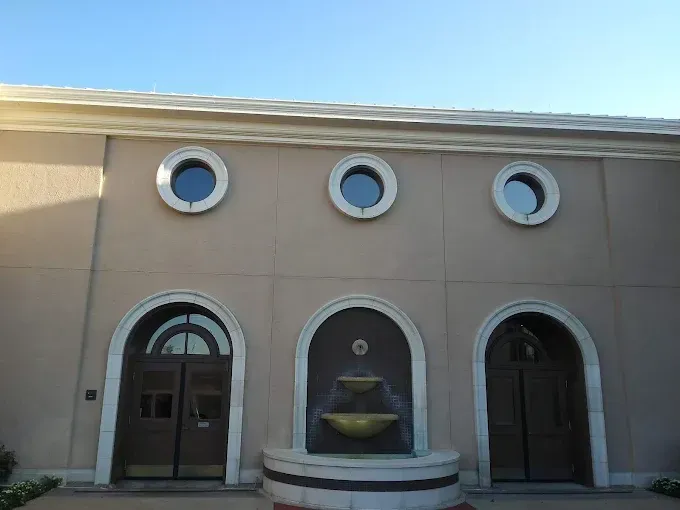 Beige building with arched doors and round windows; fountain in the middle.