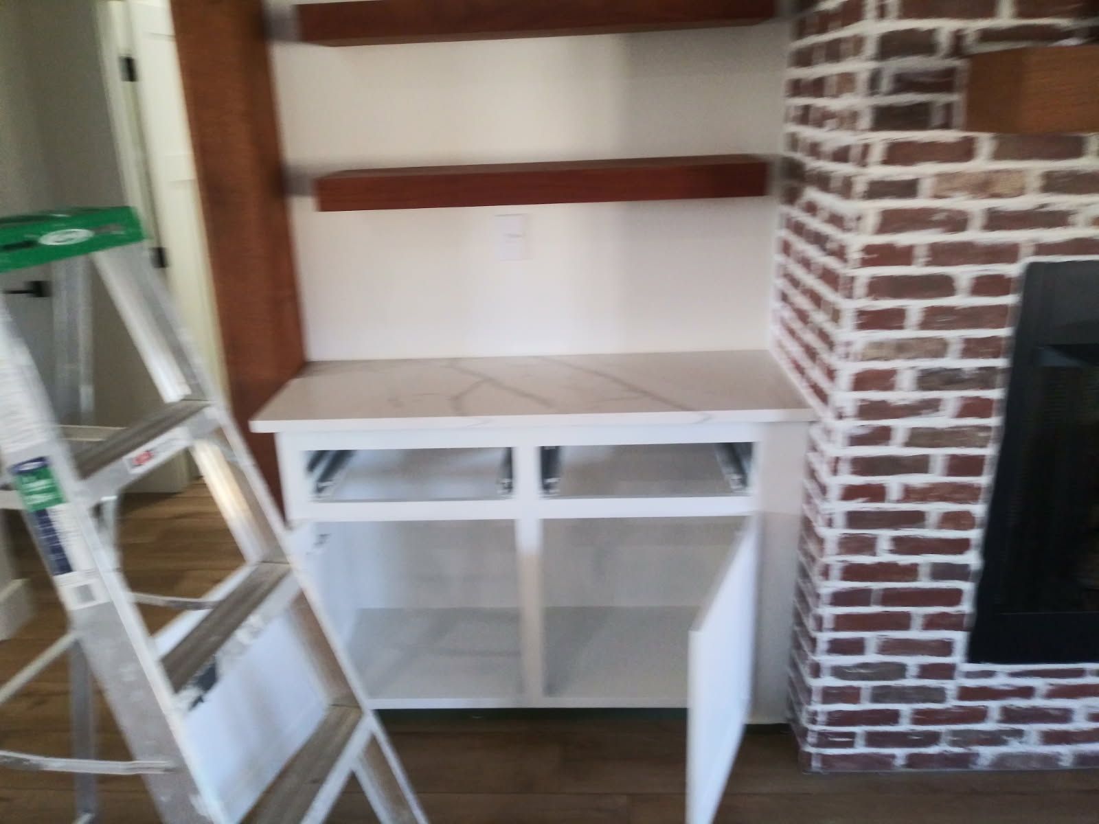 Built-in shelving and cabinets with a white countertop, next to a brick fireplace, ladder nearby.