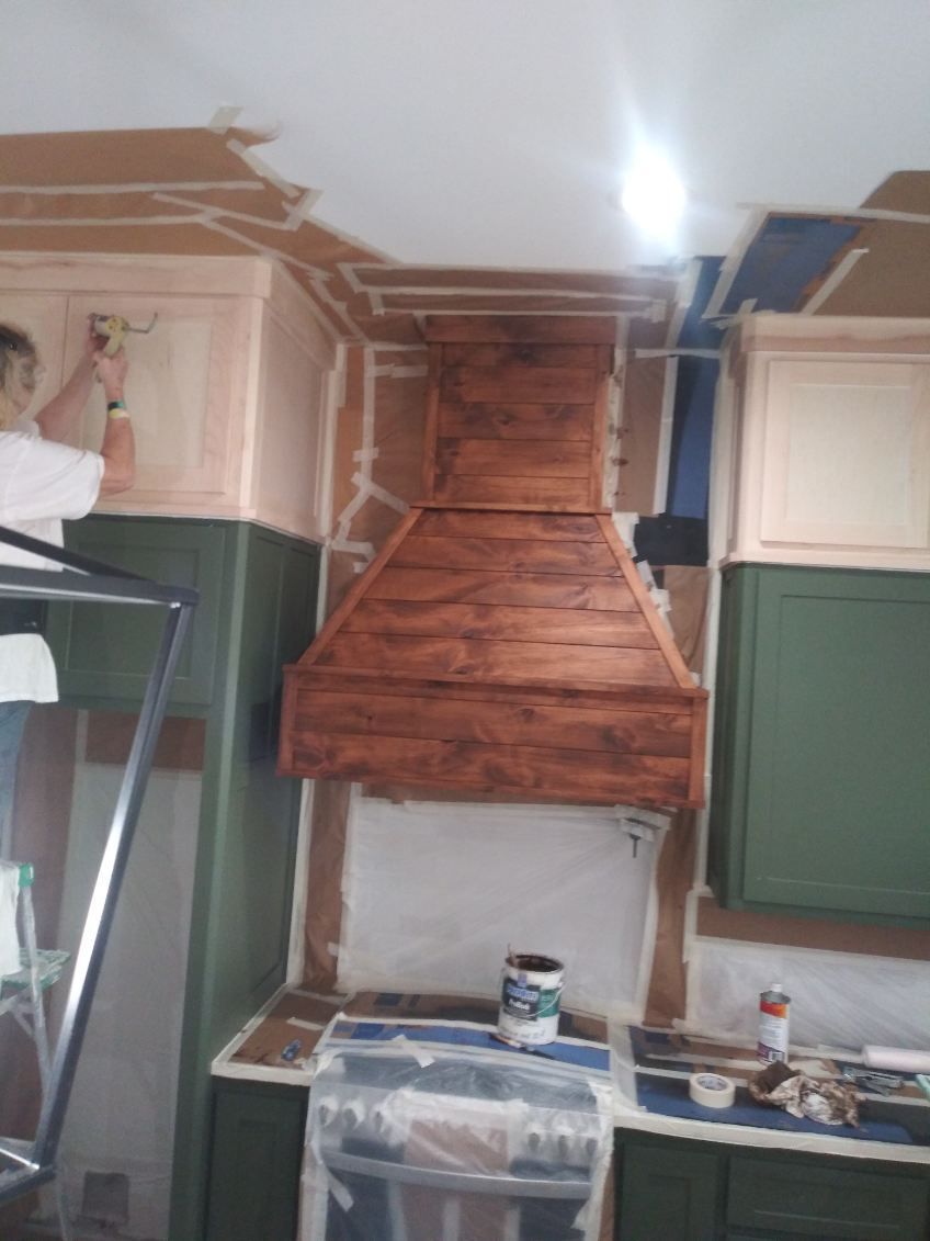Person painting a wooden kitchen hood. Kitchen cabinets and walls are taped off.