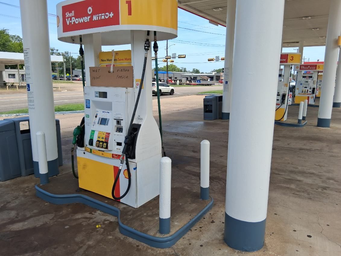 Shell gas station with pumps under a canopy. White pillars, tan pumps, and a sunny outdoor setting.