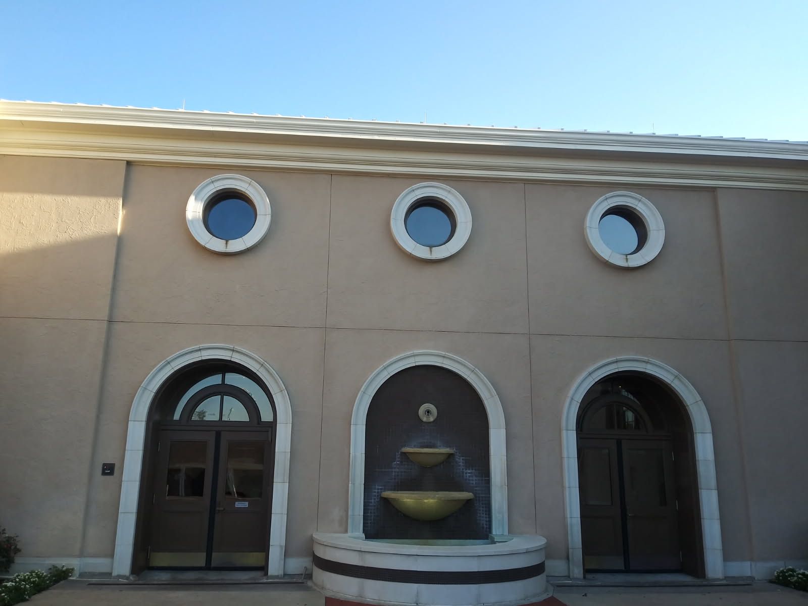 Tan building with three arched doorways, circular windows, and a fountain.