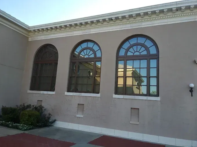 Three arched windows with dark frames in a tan building; shrubs in front.