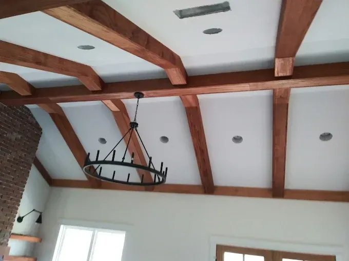 Wooden beams cross a white ceiling, with a chandelier, in a room with a brick fireplace.